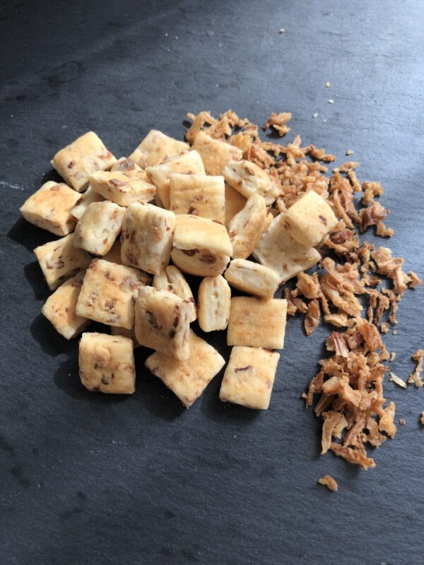 De très petits biscuits carrés déposés en tas à coté d'oignons frits, sur une ardoise