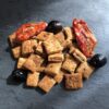 Très petits biscuits carrés déposés en tas avec deux tomates séchées et trois olives noires sur une ardoise.