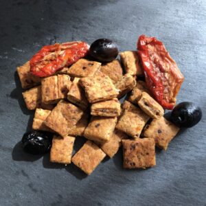 Très petits biscuits carrés déposés en tas avec deux tomates séchées et trois olives noires sur une ardoise.