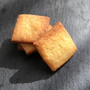 trois petits biscuits carrés au beurre, joliment dorés sont empilés en quinconce sur une ardoise. Un quatrième biscuit est posé à la verticale en appui sur la pile de biscuits
